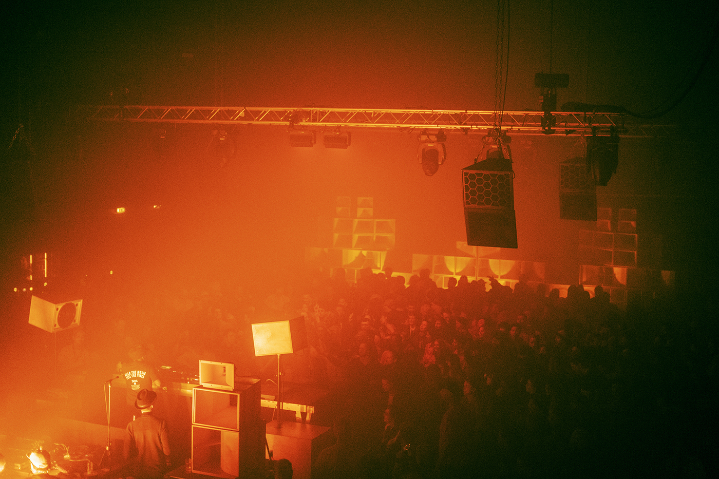 A photo with an orange glow. On the left, you can see the back of the DJ booth. On the right, you can see the room filled with people. In the back, you can see a large sound system.